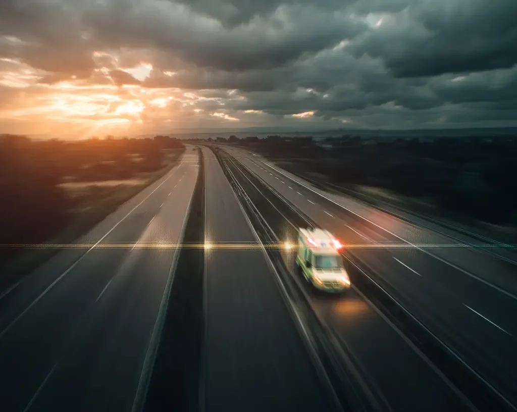 An ambulance with flashing lights speeds down an empty highway at sunset, under dramatic, cloudy skies. The road is wet, reflecting the light, creating a sense of urgency and motion.